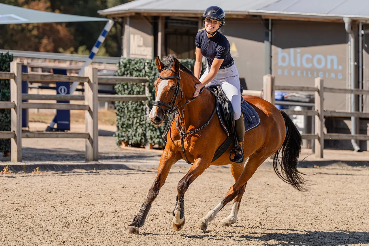 Marit van Wees galoppeert lachend op een bruin paard over een zonnige buitenpiste.