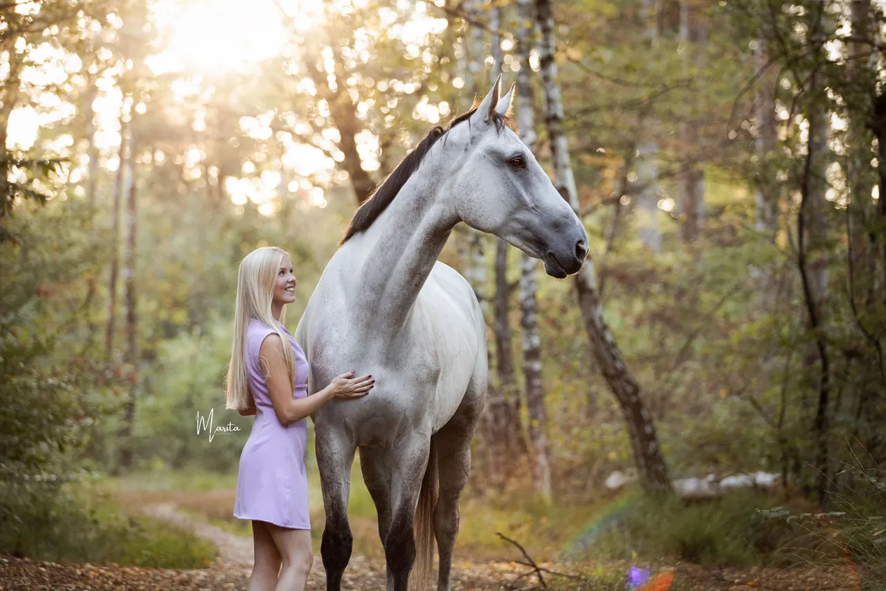 Marit van Wees staat in een zonovergoten bos naast haar paard Co en legt haar hand op de schouder van het paard.