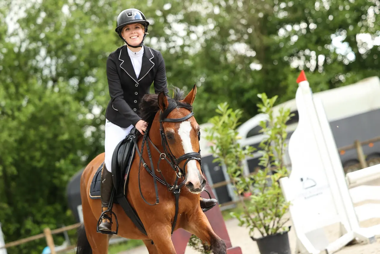 Marit van Wees lacht in de camera terwijl ze in wedstrijdkleding een bruin paard rijdt.